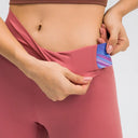Close-up of a model wearing dusty rose leggings, lifting the waistband to show a built-in side pocket with a blue and purple patterned lining.